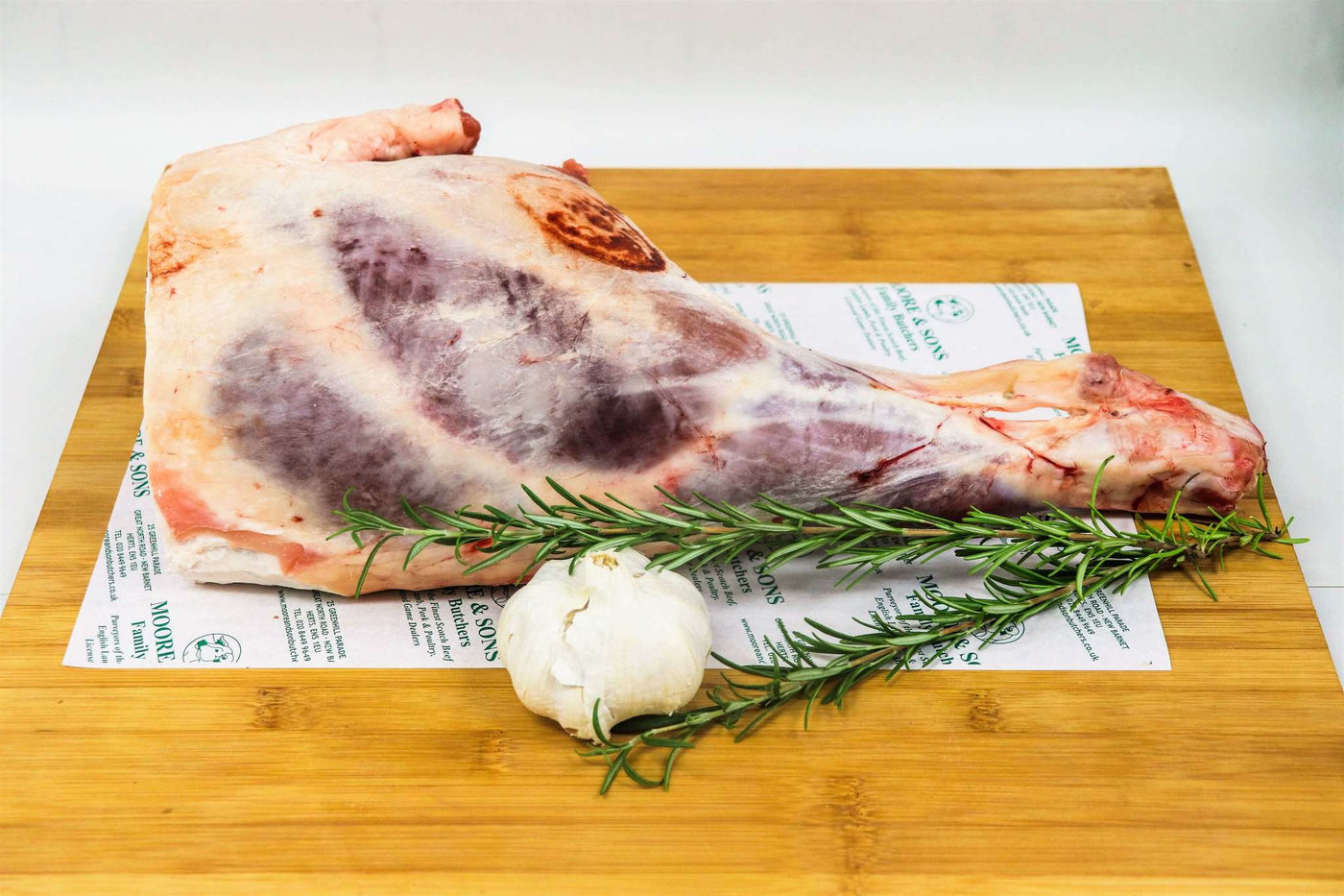 leg of lamb with bone in on a butchers block with garlic and herbs on the side