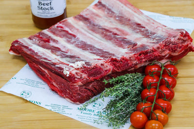 Beef short ribs on a butcher block with herbs and tomatoes on the side