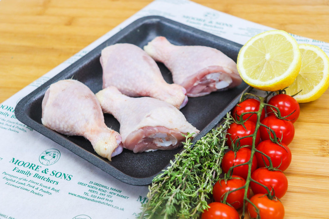 Four chicken drumstick pieces on a butcher block, with herbs, tomatoes and lemon on the side