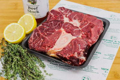 two pieces of shin of beef on a butcher block with two lemons, herbs and beef stock on the side