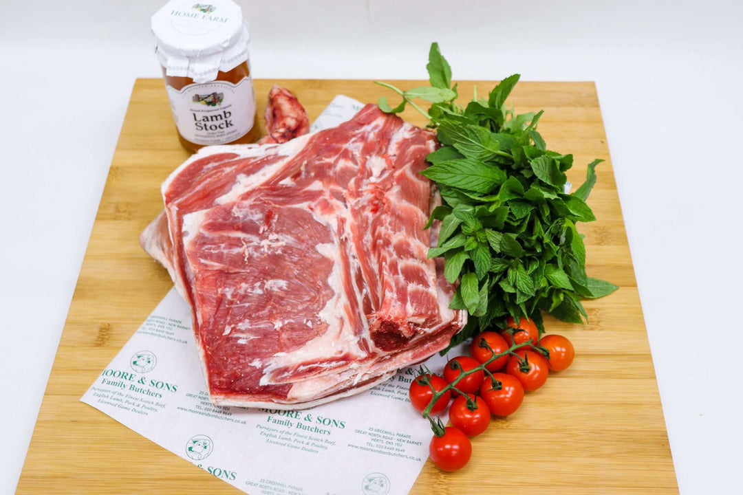 shoulder of lamb bone in on a butchers block with tomatoes, herbs and lamb stock on the side