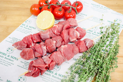 lamb skewers on a butchers block with tomatoe, lemon and herbs on the side